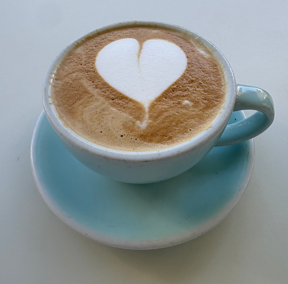 A latte topped with heart-shaped foam in a pale blue cup and saucer on an outdoor table.
